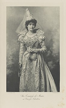 Black-and-white photograph of a standing woman richly dressed in an historical costume with a pointed hat