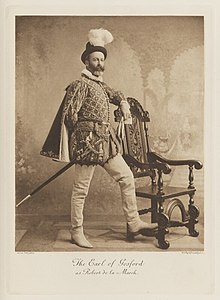 Black-and-white photograph of a man richly dressed in an historical costume with a sword, a white plume on his hat, and his foot on the rung of a chair