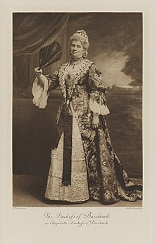 Black-and-white photograph of a standing woman richly dressed in an historical costume with a large black fan