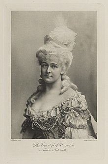 Black-and-white photograph of a bust of a woman richly dressed in an historical costume with a blonde wig, crown, and feathers