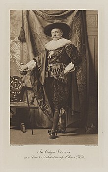 Black-and-white photograph of a standing man richly dressed in an historical costume with a large ruff around his neck, a large hat, and a sword