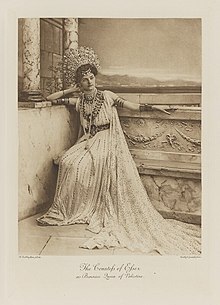 Black-and-white photograph of a seated woman richly dressed in an historical costume with a lot of jewelry and very tall jewelled crown