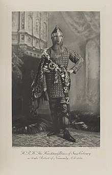 Black-and-white photograph of a standing man richly dressed in a historical costume and armor, including a large sword