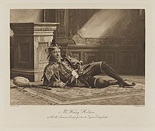 Black-and-white photograph of a man richly dressed in an historical costume of a clown and reclining on the ground