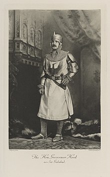 Black-and-white photograph of a standing man richly dressed in chain-mail armor, with a helmet, sword, and tabard decorated with a heraldic bird
