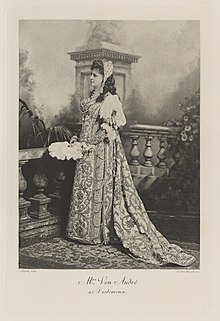 Black-and-white photograph of a standing woman richly dressed in an historical costume