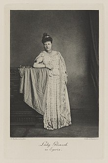 Black-and-white photograph of a woman richly dressed in an historical costume leaning on a chest