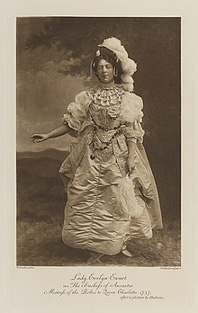 Black-and-white photograph of a standing woman richly dressed in an historical costume with feather plumes on her hat