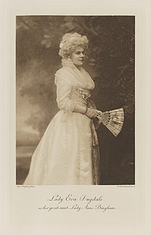 Black-and-white photograph of a standing woman richly dressed in an historical costume holding a fan