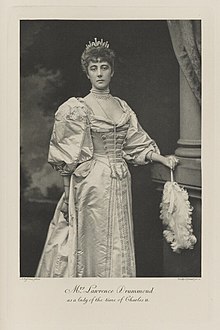 Black-and-white photograph of a standing woman richly dressed in an historical costume with a fan and a diamond tiara