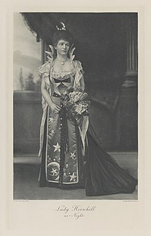 Black-and-white photograph of a standing woman richly dressed in a costume with symbols of nighttime on it and on her head