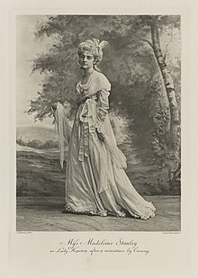 Black-and-white photograph of a standing woman richly dressed in an historical costume with a shawl in her hands