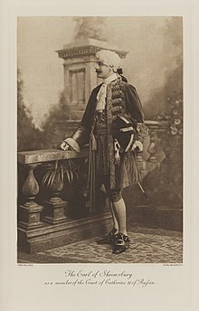 Black-and-white photograph of a standing man richly dressed in an historical costume with a white wig and large hat