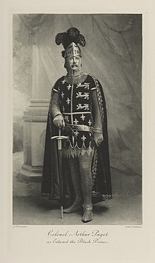 Black-and-white photograph of a standing man richly dressed in armor, with a sword, a cape and a helmet