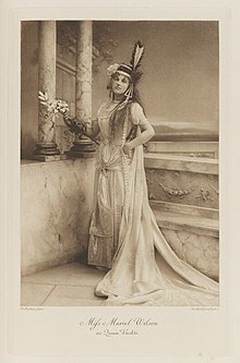 Black-and-white photograph of a standing woman richly dressed in an historical costume with flowers, big earrings, and a feather sticking up on her head