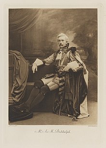Black-and-white photograph of a seated man richly dressed in an historical costume with a long cloak, a breast plate, and his feet on a pillow