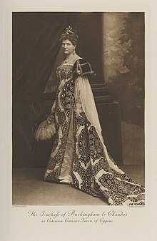 Black-and-white photograph of a standing woman richly dressed in an historical costume with a crown and a large feather fan