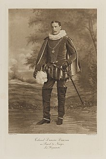 Black-and-white photograph of a standing man richly dressed in an historical costume with a ruff around his neck, a plumed hat, tall boots and spurs, and a sword