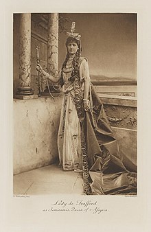 Black-and-white photograph of a standing woman richly dressed in an historical costume with a crown and a very long train