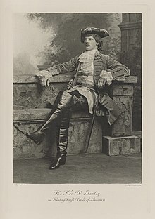 Black-and-white photograph of a sitting man richly dressed in an historical costume with boots that come up over his knees, sword, and tri-corner hat