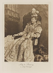 Black-and-white photograph of a seated woman richly dressed in an historical costume with a feather fan and tall feather plumes on her hat