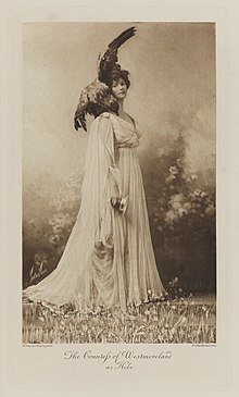 Black-and-white photograph of a standing woman richly dressed in an historical costume in a natural setting with an eagle with spread wings on her should