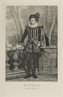 Black-and-white photograph of a standing man richly dressed in an historical costume with a short cape and a tall hat