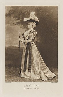 Black-and-white photograph of a standing woman richly dressed in an historical costume with a staff and very large hat