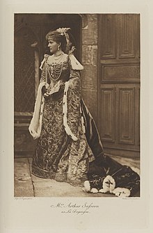 Black-and-white photograph of a standing woman richly dressed in an historical costume with lots of jewels and ermine