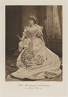 Black-and-white photograph of a seated woman richly dressed in an historical costume with a scepter and crown