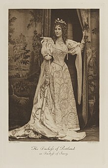 Black-and-white photograph of a standing woman richly dressed in an historical costume with a crown and several strands of pearls