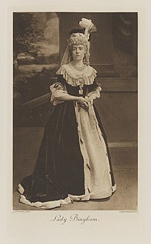 Black-and-white photograph of a standing woman richly dressed in an historical costume, with a closed fan and a high hat with a feather plume