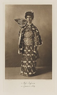 Black-and-white photograph of a standing woman richly dressed in a traditional Japanese kimono with a partially opened fan