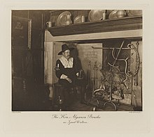 Black-and-white photograph of a man richly dressed in an historical costume sitting in a fireplace that does not have a fire and holding a tankard