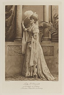 Black-and-white photograph of a standing woman with a feather fan richly dressed in an historical costume
