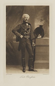 Black-and-white photograph of a standing man richly dressed in an historical military costume with tall boots and a fur hat next to him