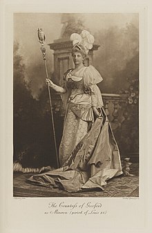 Black-and-white photograph of a standing woman richly dressed in an historical costume with 3 white feather plumes on her head and a long staff with an owl on top