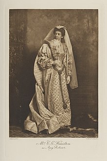 Black-and-white photograph of a standing woman richly dressed in an historical costume with a veil and fan