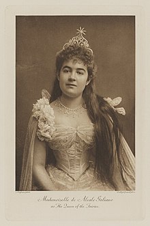 Black-and-white photograph of a woman richly dressed in an ornate costume with her hair down and a crown with a star on top