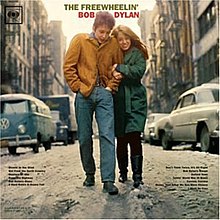 The Freewheelin' Bob Dylan's album cover. Wearing a brown jacket and blue jeans, a man walks along a snowy street. A woman wearing a long green coat and black pants holds onto his arm and walks alongside him. The words "The Freewheelin' Bob Dylan" frame the man's head, and the names of songs contained within the album are listed in small print in the bottom left and right of the image.