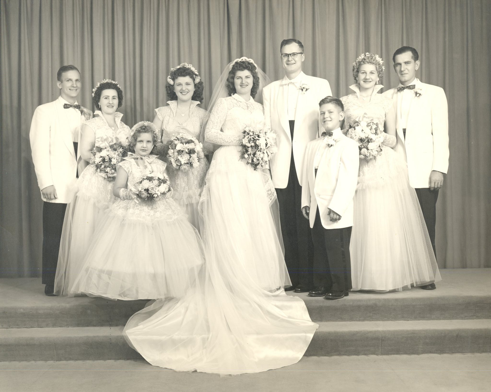 Black and white photograph of a wedding party. The bride's dress has a long train. The three bridesmaids and flower girl are wearing dresses with full gauzy skirts and standing collars.