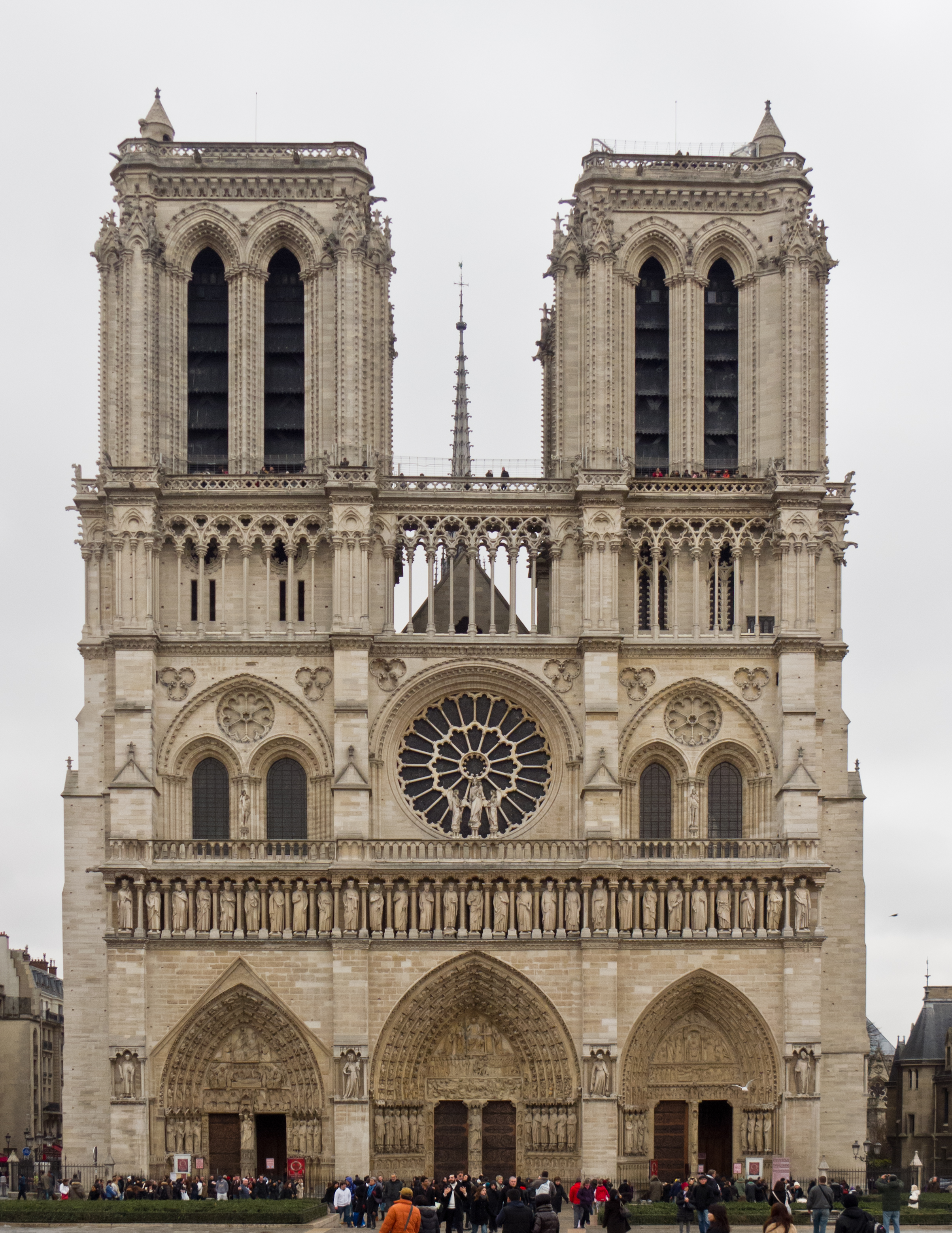 Photo of Notre Dame from the outside. Two towers sit side by side with various ornate architectural details. There are three arched doorways along the ground, the second row has a circle center, with two side by side arches on either side, as well as a pointed tower in the center. 