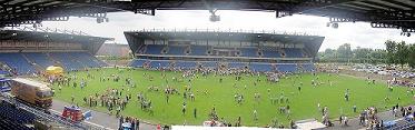 A view of a football stadium with a stand opposite and to the left, and an open space to the right. There are people on the pitch