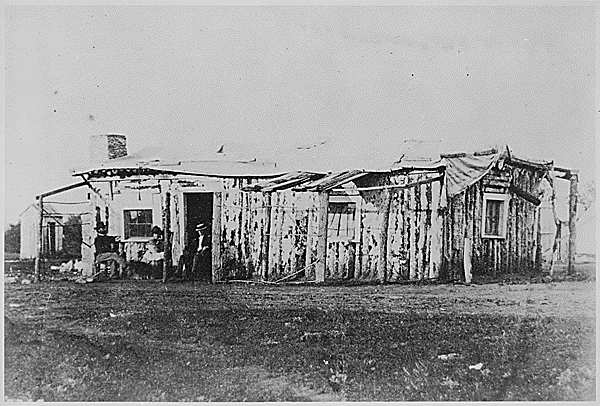 File:"Officers quarters at Ft. Rawlins, Wyoming, May 7, 1877." Two men and a woman holding a baby sit in front of the crude structure - NARA - 531107.gif