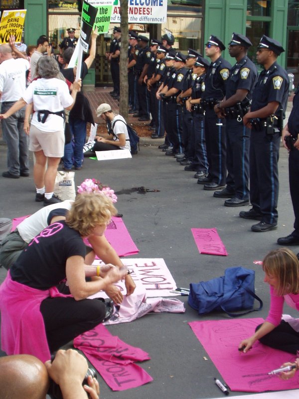 Photo of a line of Boston police facing Code Pink activists preparing signs during an anti-war demonstration in Boston during the 2004 Democratic National Convention