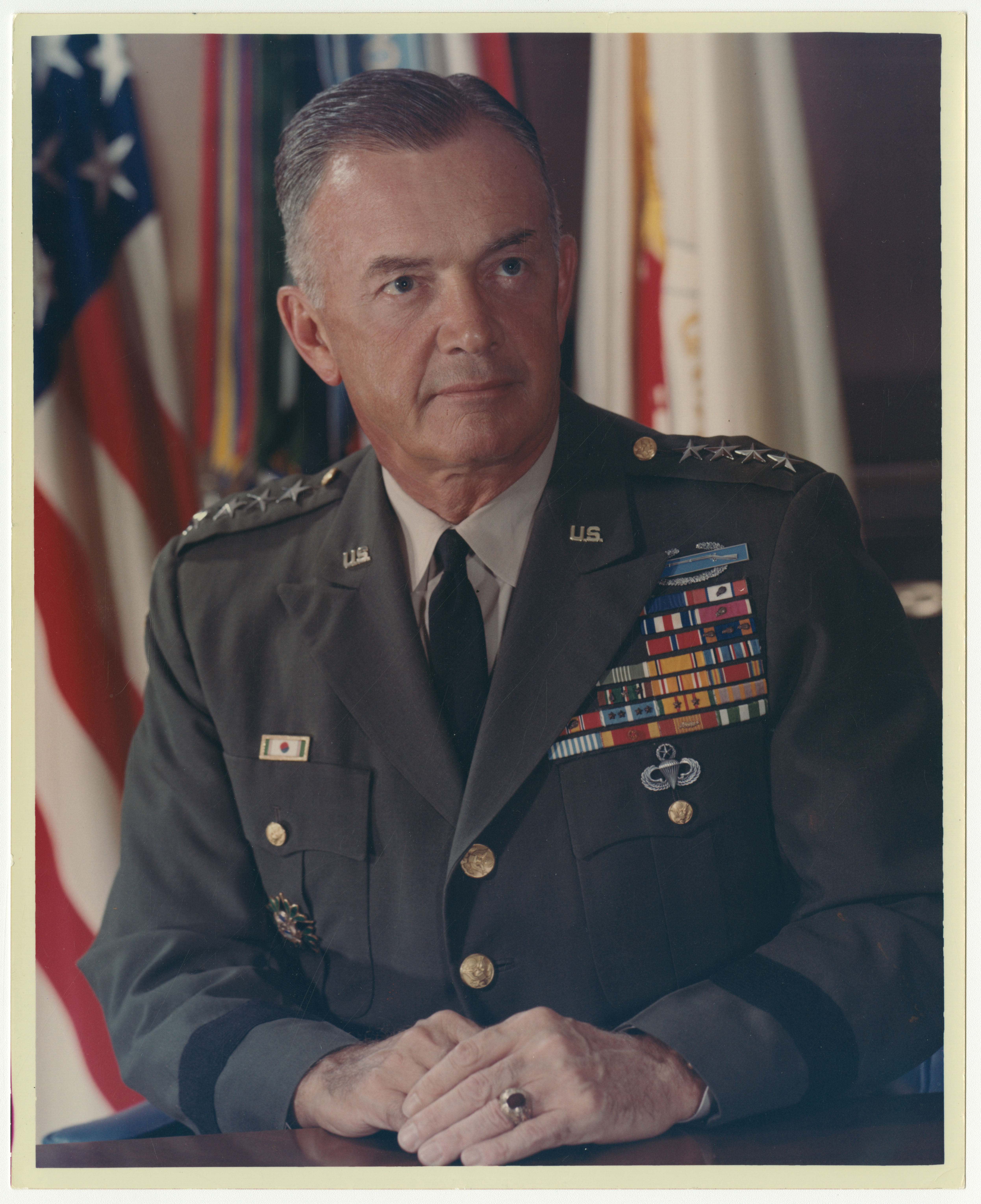 White-haired, clean-shaven man in dark-green ceremonial military uniform and tie. He has a lot of insignia on the left side of his chest and stars to indicate that he is a general.