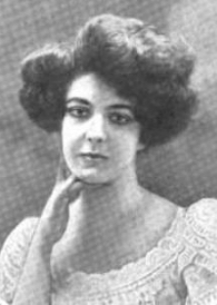 A white woman with bouffant dark hair, with her hand on her chin; she is wearing a white lacy dress with a round neckline.
