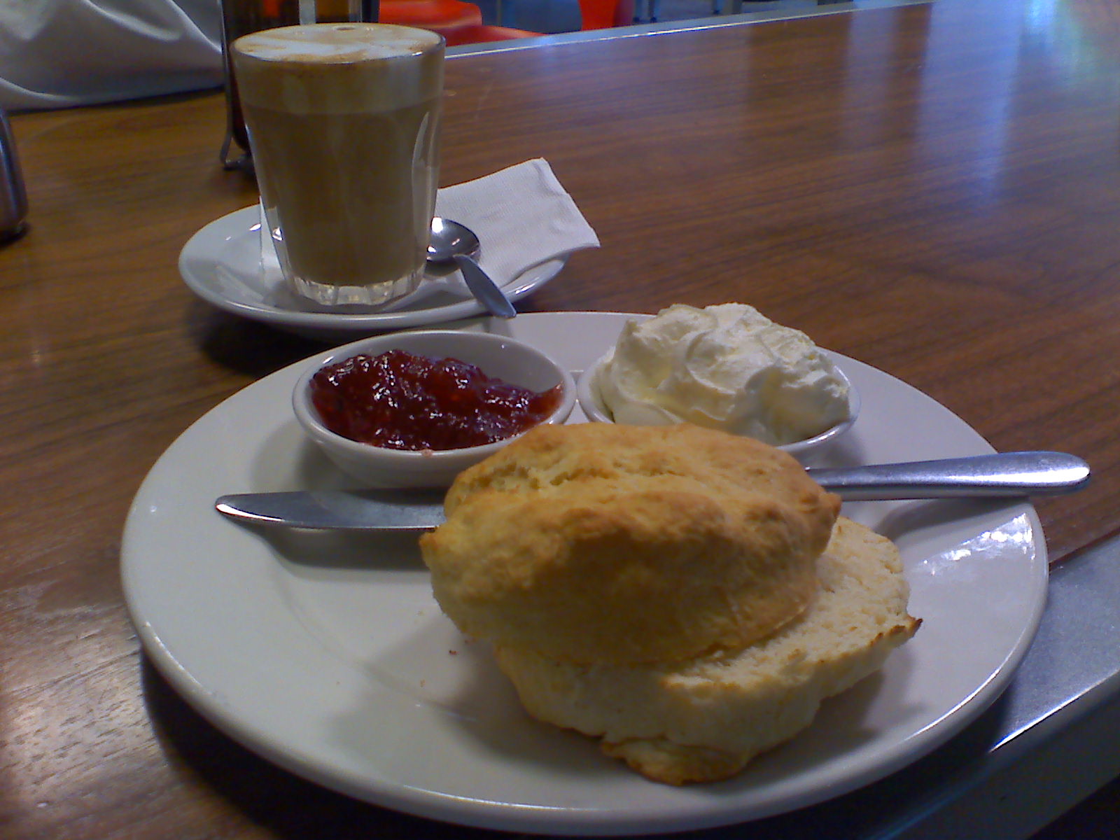 A white plate laden with a scone and a dish of jam and a dish of clotted cream with a glass cup filled with milky tea on a wooden tabletop.