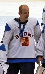 Een blanke ijshockeyspeler gekleed in een blauwe trui met groene en witte bies, evenals een blauwe helm.  Hij staat ontspannen op het ijs, met zijn hockeystick in één hand.