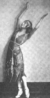 A white woman in a standing dance pose, arms raised over her head, wearing a dress with a plunging V-neckline and a tassled cord belt